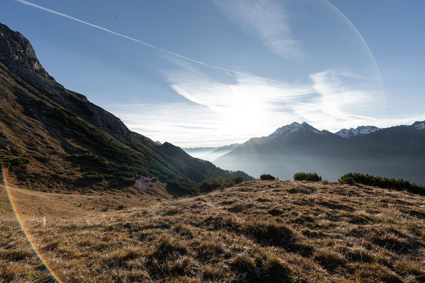 Bergwiese bei Sonnenaufgang mit Sonne hinter dem Gipfel und Nebel im Tal, Arlberg