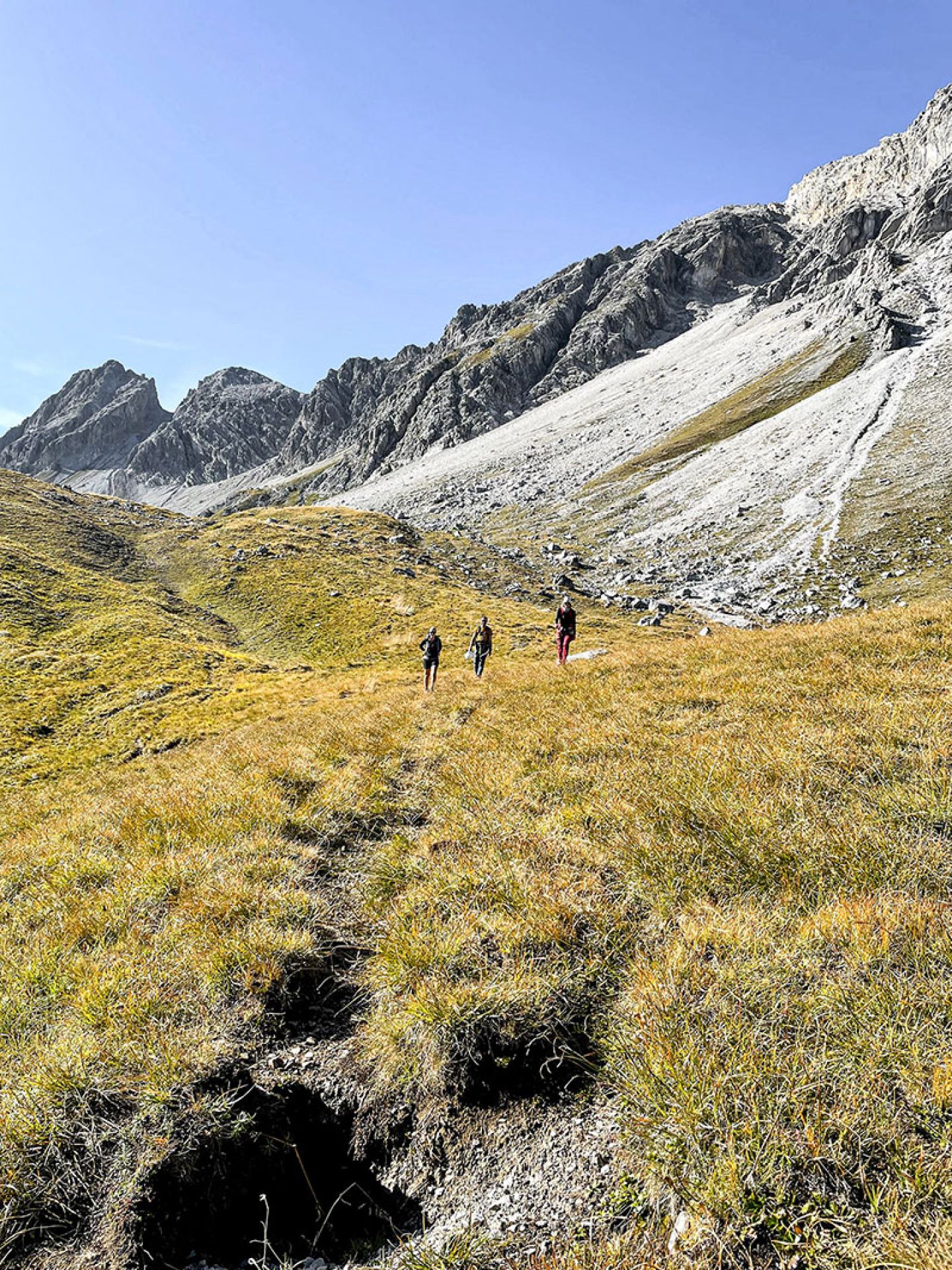 Three hikers crossing an alpine meadow towards the rocky peaks on the Arlberg