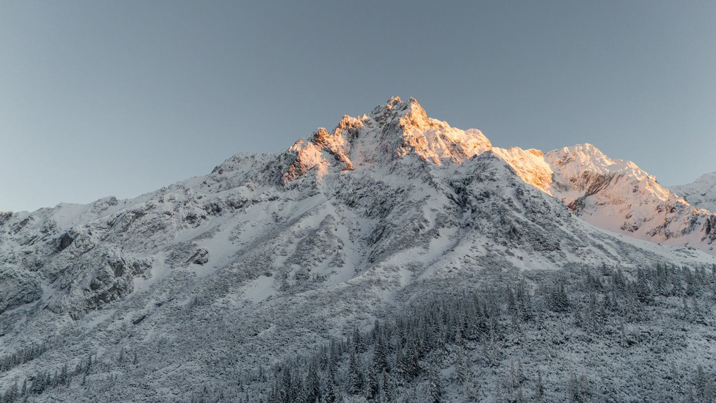 Drohnenaufnahme: verschneiter Gipfel mit Alpenglühen über bewaldetem Hang im Winter am Arlberg