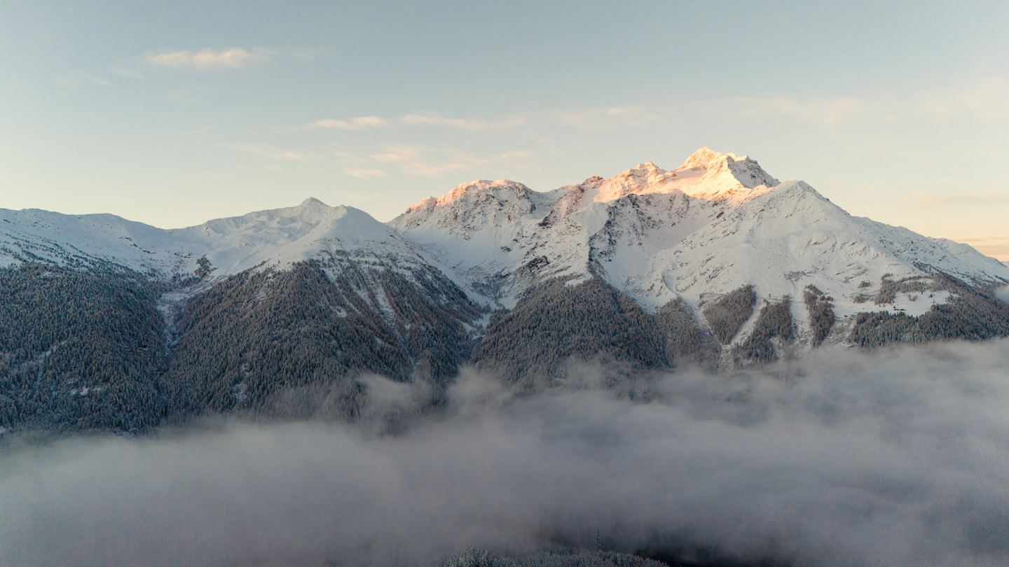 Drohnenaufnahme: verschneiter Gipfel mit Alpenglühen über Wald und Nebelmeer am Arlberg