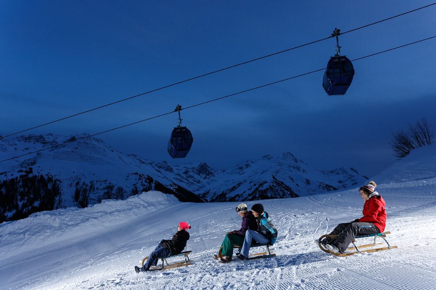 Familie beim Rodeln in der Dämmerung unter den Gondeln der Arlberger Bergbahnen